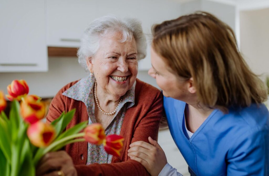 An older woman arranging tulips in a vase and smiling at a female nurse who is holding her arm and looking at her.