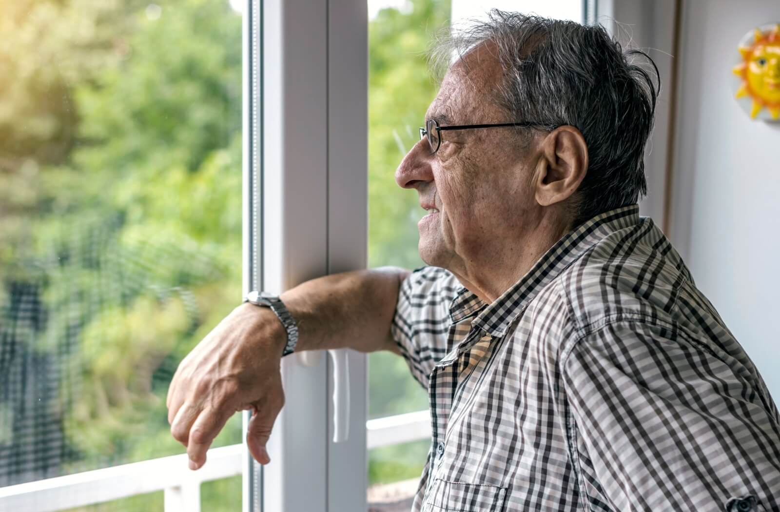 An older adult man leaning into a window with his right elbow and looking out of the window with a serious expression.