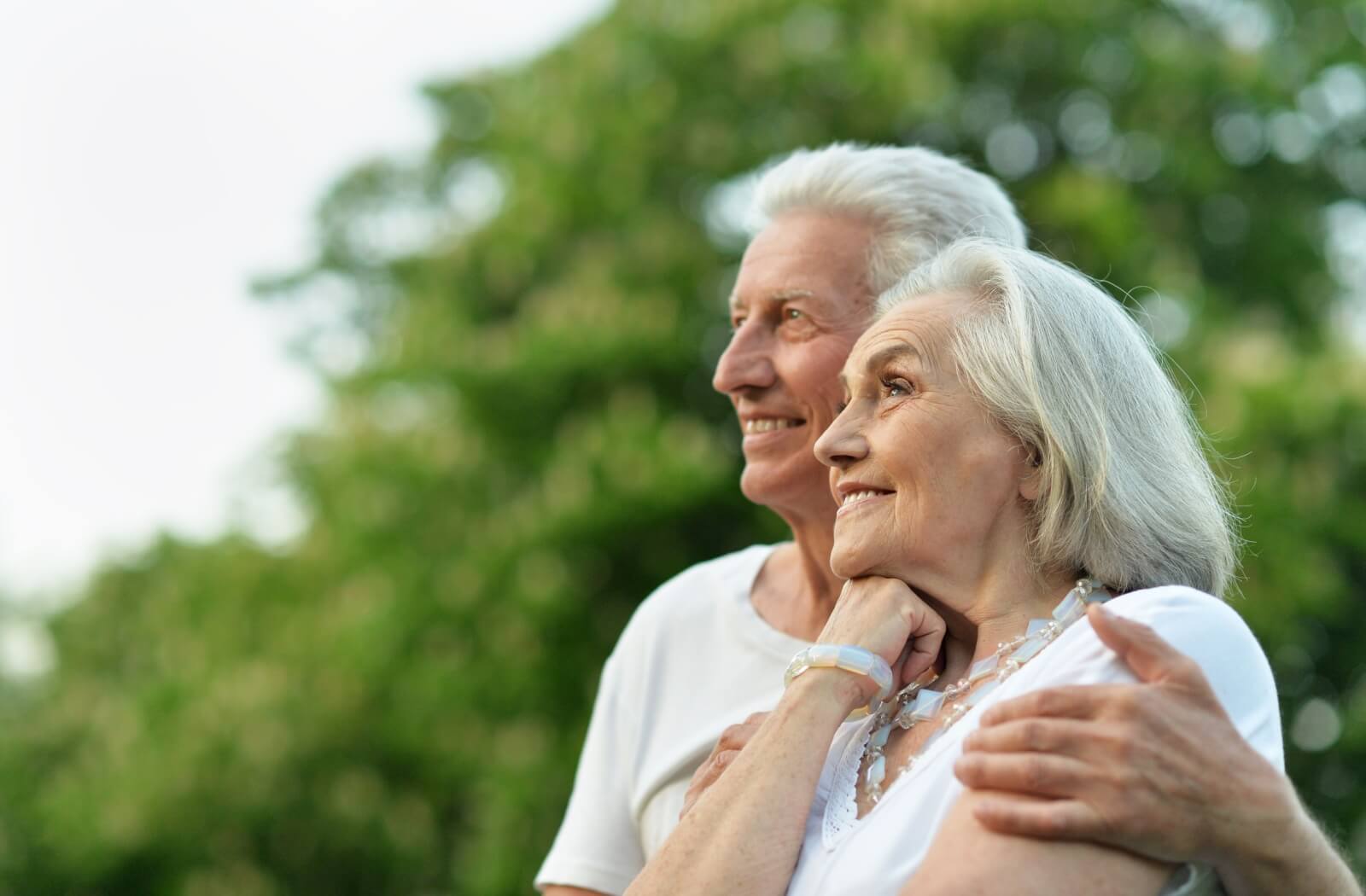 A side view of an older man with his arm wrapped around an older woman while they smile into the distance.