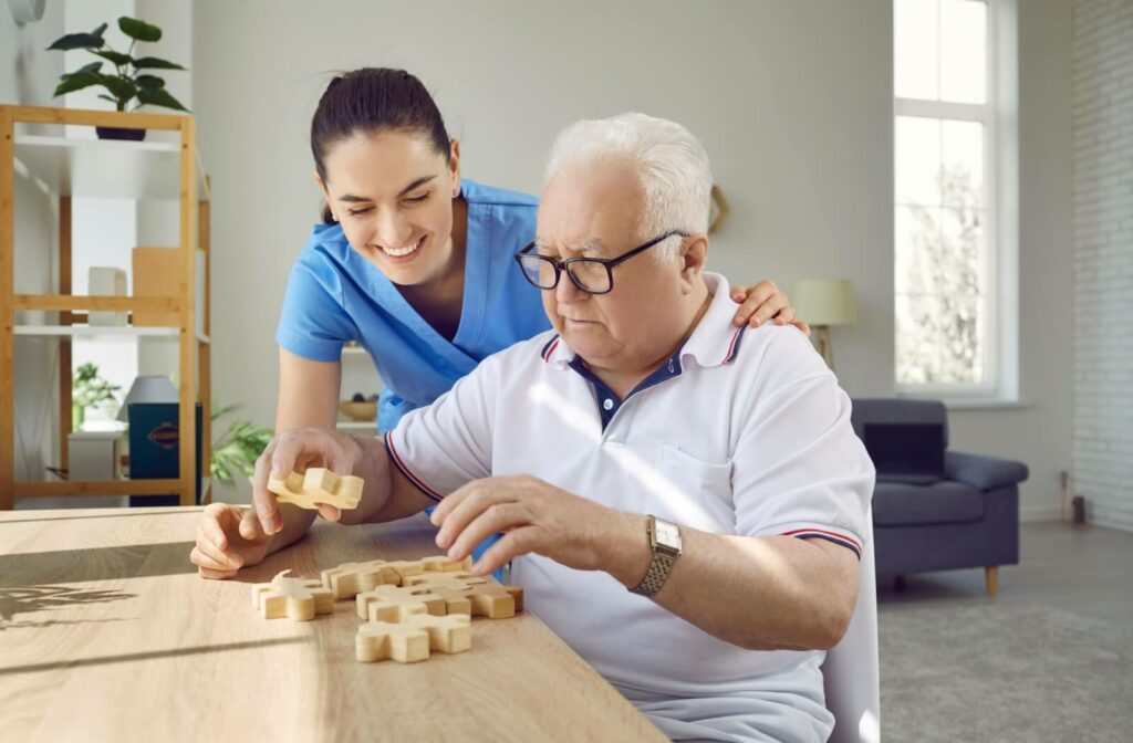 An older man with glasses sitting at a table and doing a jigsaw puzzle with the help of a caregiver.