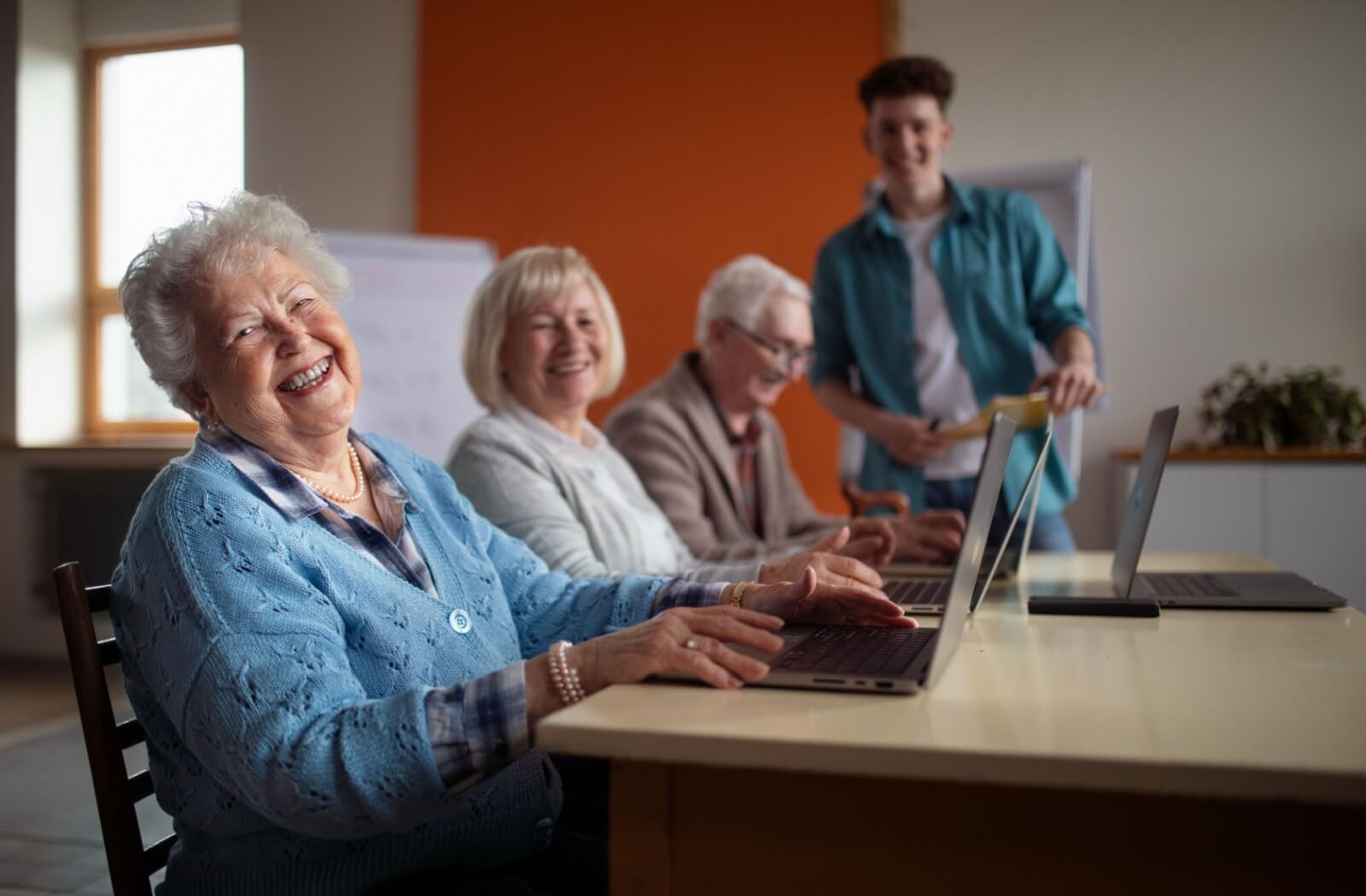 Three smiling seniors sit in front of laptops, being taught tech tips by a young adult at a senior living community