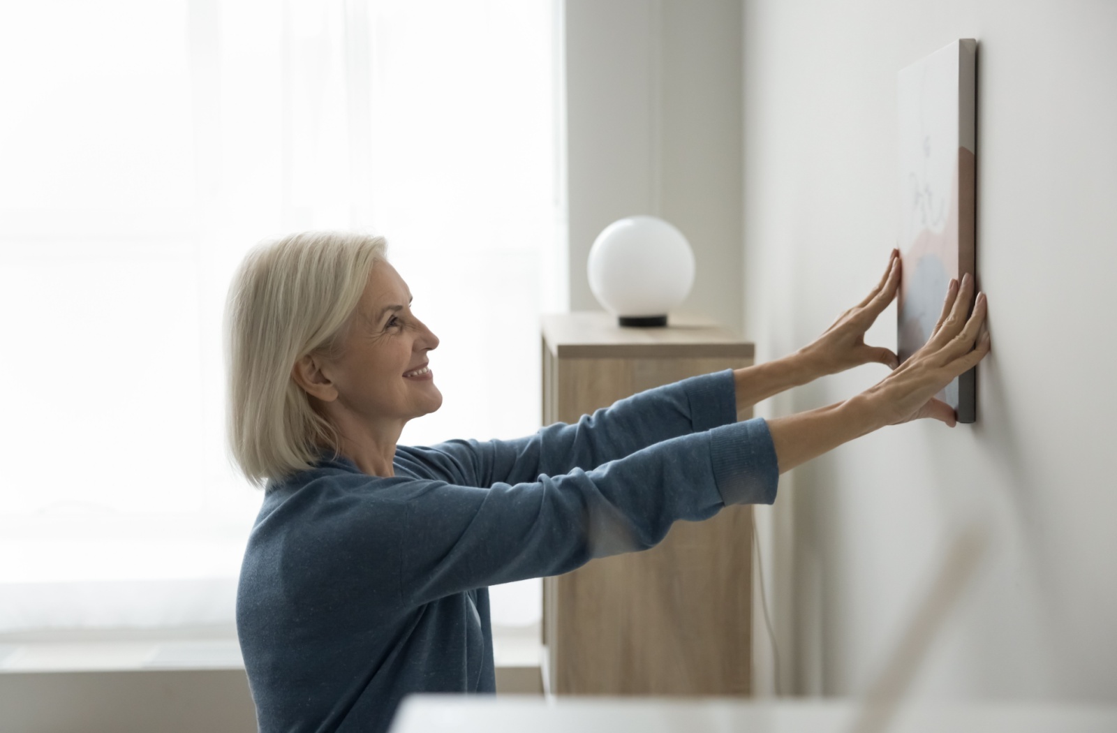 An older adult hangs up a picture in their assisted living room.