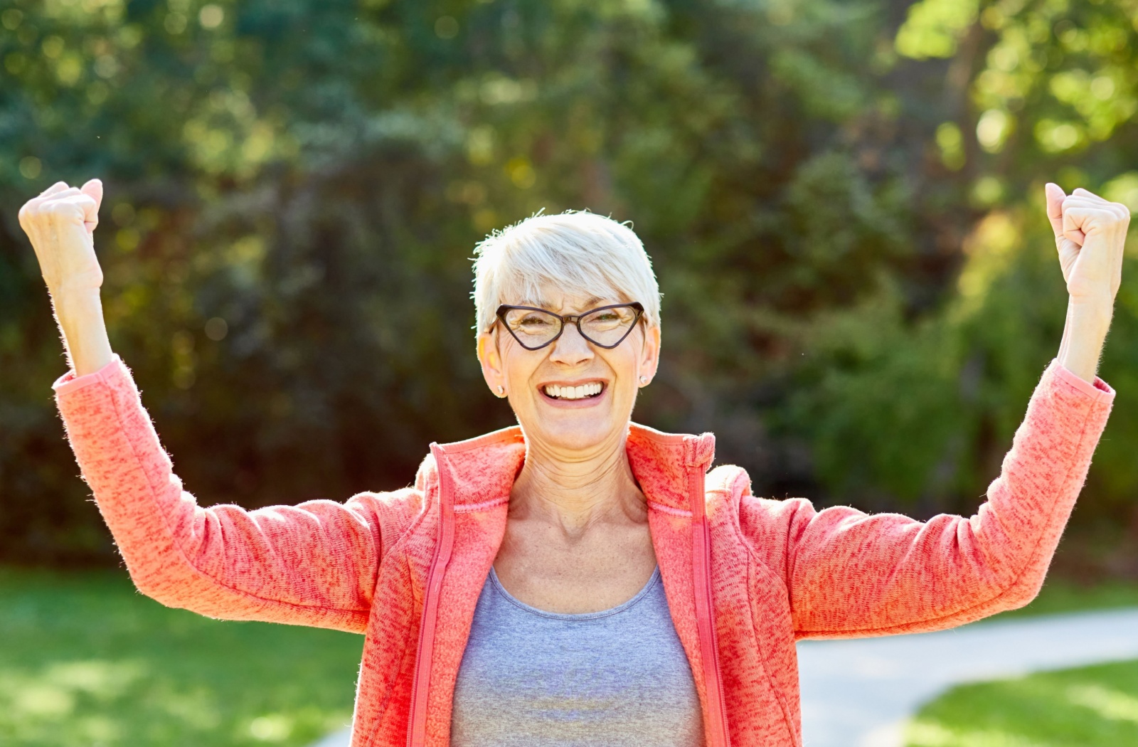 Smiling older adult in athletic clothing raising arms outdoors on a sunny day in a green park setting.