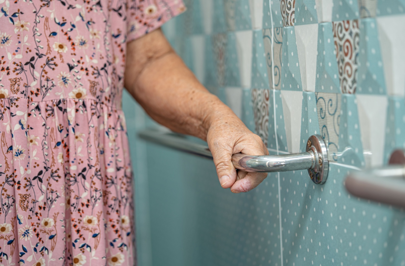 Older adult holding onto a wall-mounted grab bar in a tiled shower for stability and support during bathing.