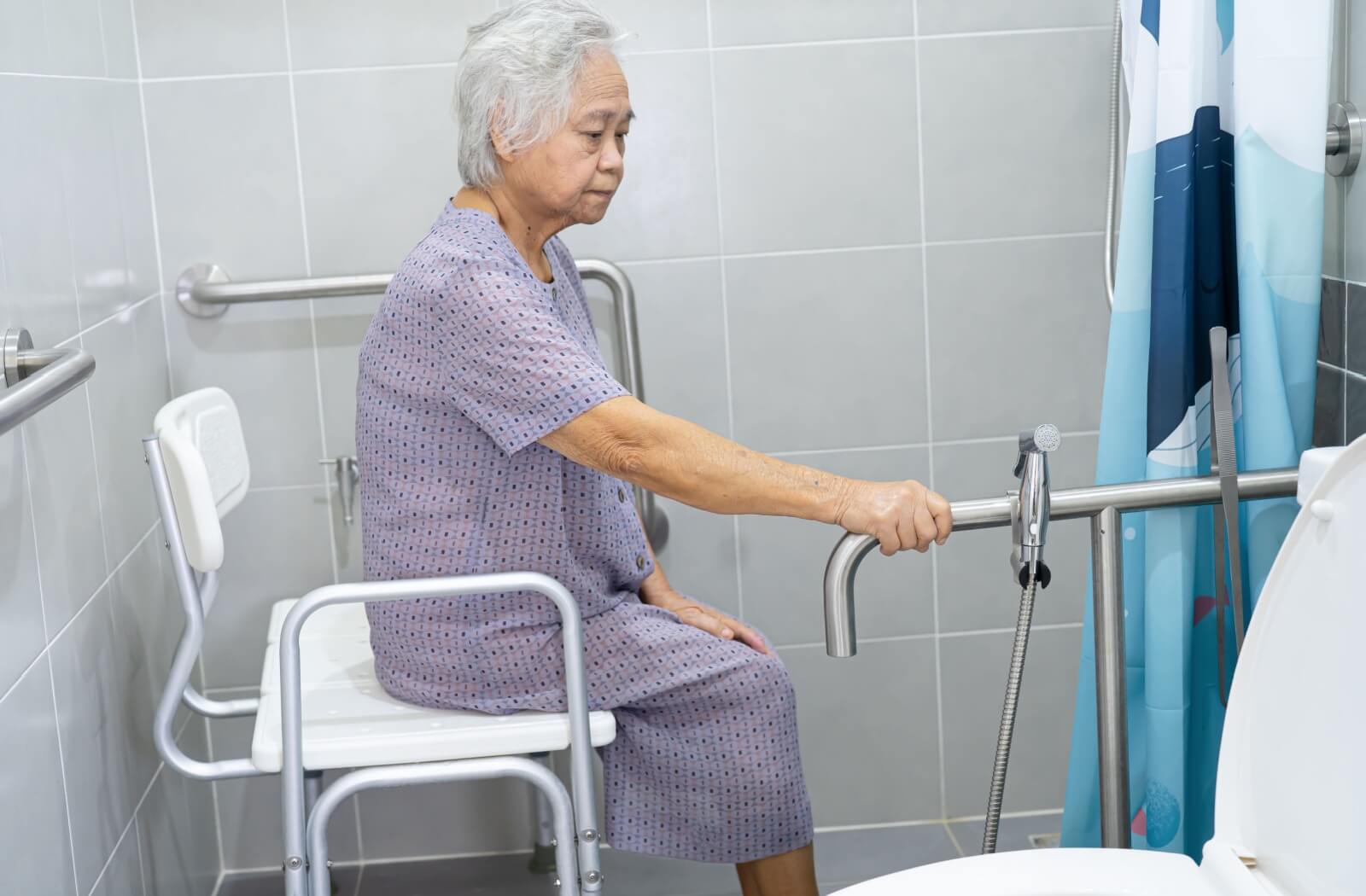 Older adult sitting on a shower chair holding a grab bar in an accessible bathroom with safety rails and handheld sprayer.