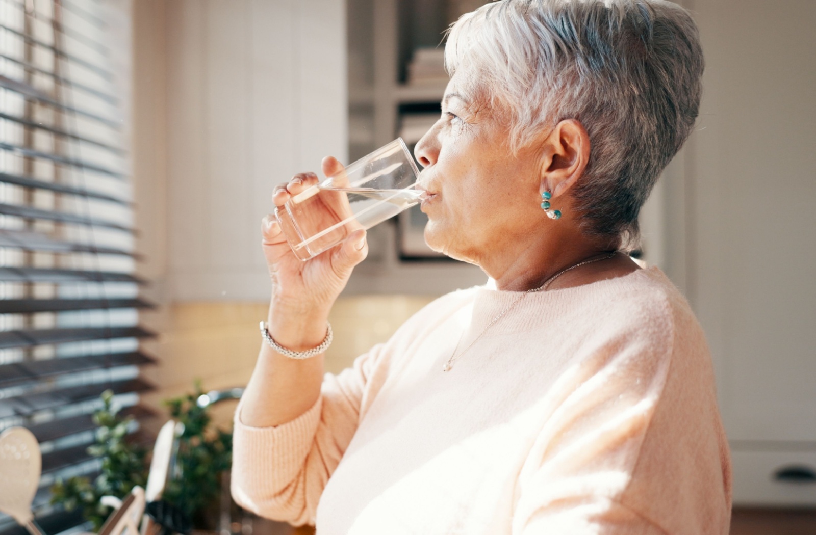 Older adult standing by a window drinking a glass of water to stay cool and hydrated in warm weather.