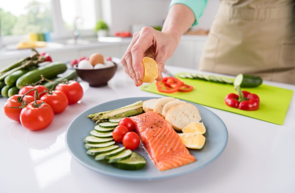 A senior woman squeezing lemon on a filet of salmon on a plate with some cherry tomatoes, sliced cucumbers and asparagus.