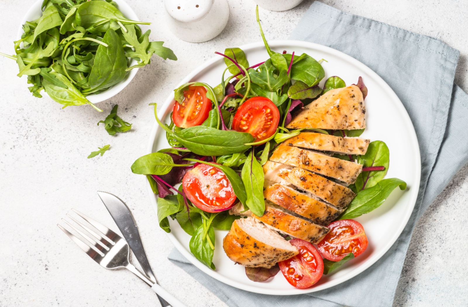 A plate of well cooked chicken breast with tomatoes, basil and kale set on a white table.
