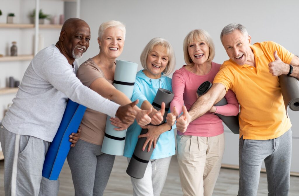 A group of seniors with yoga mats all smile and give a shared thumbs up after an enjoyable yoga class together