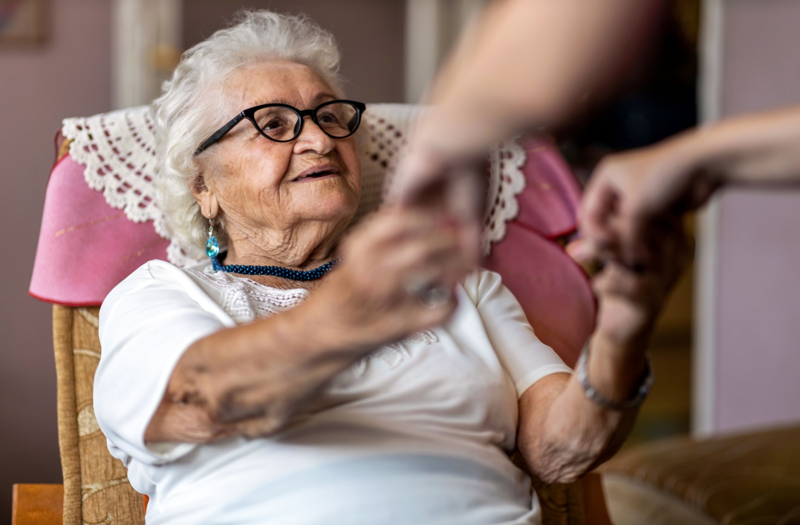 An elderly woman holding hands with a caregiver while smiling at them, sitting in a chair.